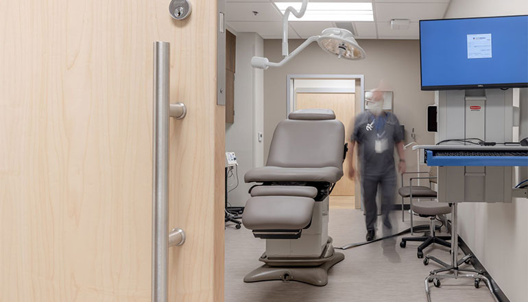Medical professional standing in an exam room. A sliding door pull handle is shown in the foreground of the photo. 
